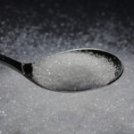 A metal spoon holding processed white table sugar over a dark background